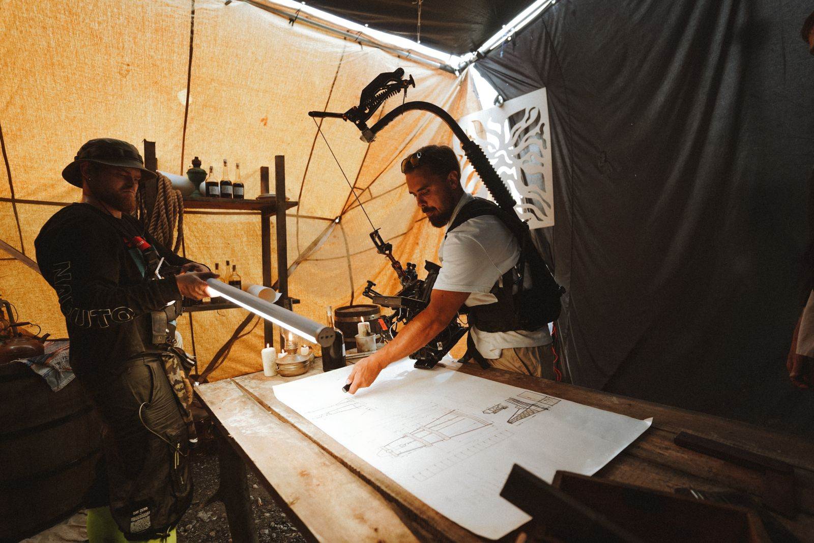 Two engineers use detailed plans inside a tented workshop, focusing on construction or design projects