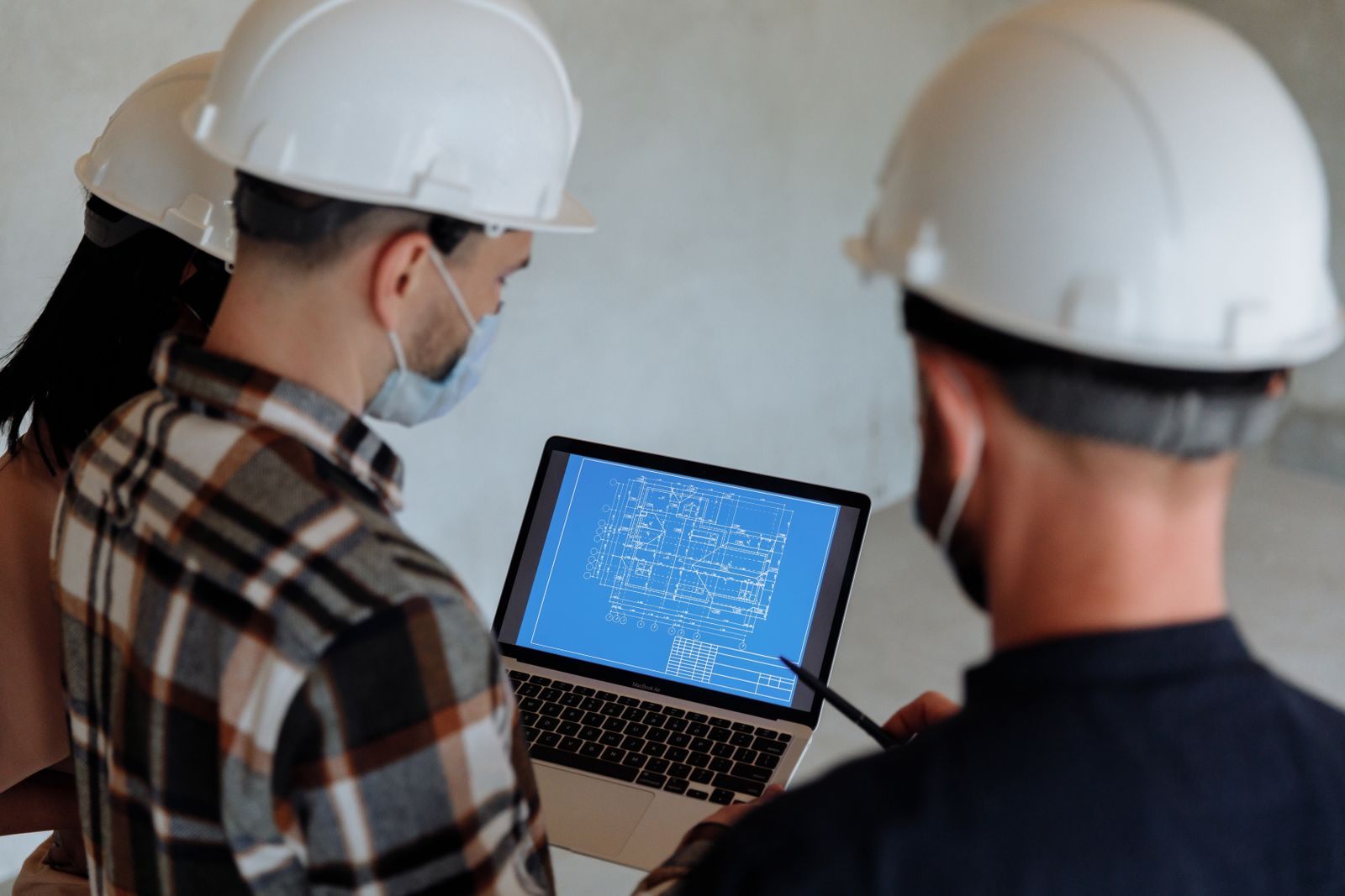 Engineers wearing hard hats and masks review a construction blueprint on a laptop indoors